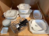 Overhead view showing 4 CorningWare Cornflower casserole dishes with lids in various sizes, two white open oval French White bowls, several clear glass bowls, small rectangular white dishes, and metal holders with black handles.