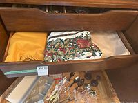 Photo showing table linens folded in a drawer including yellow, floral, and beige cloths; glass candlestick holders and other dining accessories seen beneath drawer