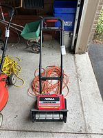 Front view of the Noma TurboThrower snow blower with orange extension cord coiled behind it, showcasing the handle and main body.