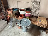 Photo showing assorted plant pots in multiple sizes and colors, wire tomato cages stacked vertically, and a grey bird bath on the floor along with folded cardboard leaf bags.