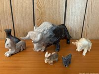 Six carved animal figures arranged on a wooden surface with wooden panel background: two bulls (one large black/grey, one smaller white), two small donkeys, a marble cat, and a small carved stone cat.