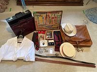 Photo of all items laid out on carpet: white suit jacket, brown leather garment bag, multiple briefcases, ties, belts, hats, framed swizzle sticks, and accessories