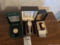 Three men's watches displayed side by side in their original or appropriate boxes on wooden surface.