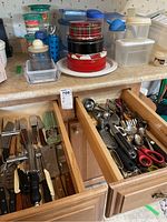 Kitchen countertop with stacked tins and various plastic storage containers with lids; open drawers revealing kitchen knives, peelers, graters, measuring spoons, and scissors.