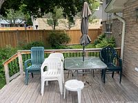 Photo showing four plastic resin stackable chairs in three styles, glass patio table, neutral taupe umbrella, and small white resin side table on wooden deck.