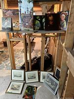 Wide shot of multiple framed prints leaning against wall and paintings arranged on makeshift shelf, showing variety of items in the lot.