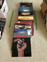 Photo showing a vertical stack of 11 vinyl record albums on carpet, featuring The Kinks, Billy Joel, Saturday Night Fever soundtrack, Placido Domingo and more.