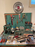 Photo of a workbench area showing screwdrivers, files, hammer, wrenches, pliers, and miscellaneous hand tools hanging on a green pegboard with additional tools and parts on the work surface.
