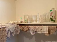 Glass vases lined up on a shelf with crocheted doilies draped below, showing the mix of glass and fabric items in the lot.