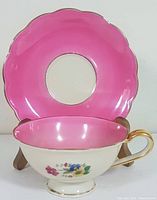 Front view of teacup showing floral design, pink interior, and saucer with scalloped bright pink top and gold trim.