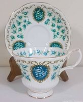 Front view of teacup with saucer displayed on wooden stand. Shows detailed floral cameo medallions in teal and gold on white fine bone china.