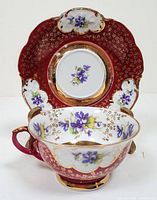 Teacup and saucer set displayed frontally showing detailed purple floral design, gold accents on burgundy background, crown-shaped scalloped edges