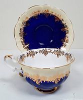 Tea cup and saucer front view showing cobalt blue, white, and gold floral decorative pattern with scalloped edges.