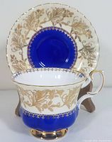 Front view of teacup with saucer in background showing detailed gold floral design with cobalt blue band and gold trim