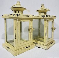 Front and angled view of two rustic metal and glass lanterns with distressed cream paint and black accents, showing square bases, glass panels, metal doors with latches, and hanging rings on top.
