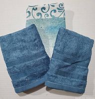 Two solid teal towels with ribbed texture bands in front of a teal and white patterned towel.