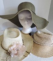 Front view of three sun hats including a large olive green wide brim hat on mannequin and two smaller straw hats in front: beige with pink flower and light brown woven hat.