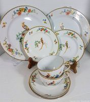 Front view of 2 dinner plates, 2 bread plates, and 2 stacked cups and saucers showing golden pheasant and floral pattern with gold trim.