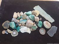 Top-down photo of a collection of mineral specimens on black background. Includes chrysocolla, rose quartz, geode slices, petrified wood slab, and various other stones.