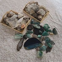 Two wooden trays containing rough mineral specimens mostly whitish and light brown, alongside scattered polished and rough stones including greenish chrysocholla, blue banded geode slices, pink rose quartz pieces on a patterned fabric surface.