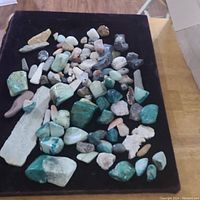 Photo of mixed mineral and rock specimens on a black surface showing Chrysocholla, quartz, petrified wood, black tourmaline, and volcanic glass stones.