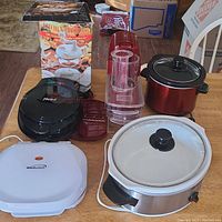 Photo of small kitchen appliances including two Crock Pots, an omelet maker, grilled cheese maker, and a boxed Kitchen Plus 2000 food processor.