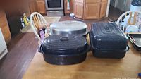 Three large turkey baking pans on a kitchen table: one large aluminum pan with lid and two black speckled enamelware pans with lids.