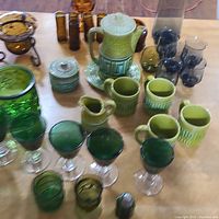 Photo of entire glassware assortment including green ceramic teapot set with cups, green glass goblets and tumblers, amber and blue shot glasses, and clear glass pitcher.