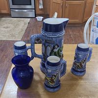 Box containing large ceramic beer pitcher and three matching ceramic beer steins with raised decorative figures, along with a cobalt blue glass vase, placed on wooden table.