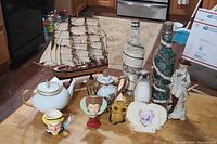 Photo shows a collection of vintage boat-themed teapots, a wooden model ship, decorative bottles, and multiple figurines arranged on a floor in a kitchen area.