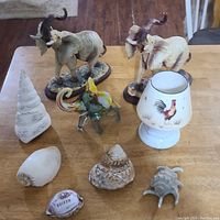 Photo showing three vintage elephant figurines, multiple decorative sea shells, and a small lamp with a rooster design on a wooden table.