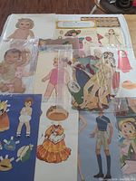 Photo of multiple vintage paper dolls and paper clothes laid out on floor showing various baby and child figures alongside a multi-piece snap-together paper puppet set in packaging.