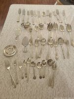 Overall layout of vintage silver and silver plate spoons, forks, and additional pieces on white textured cloth showing number, condition, and assortment.