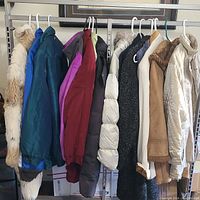 Photo showing a row of women's coats and vests hanging on a rack with assorted colors and styles, including puffer jackets, faux fur, quilted and shearling materials.