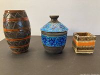 Three ceramic pieces on black surface: a tall brown and orange geometric vase, a blue lidded vessel with geometric details, and a small square ashtray with chips to rim.