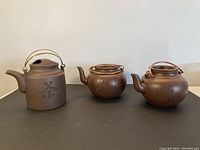 Front view of all three clay tea pots arranged side by side, showing the variation in shape and metal handles