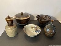 Five studio pottery pieces displayed side by side against a neutral wall. Includes a lidded pot, pitcher, bowls, and a bottle.