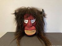Front view of the carved Iroquois False Face mask showing red face with large white eyes, wooden carved teeth, two small horns, and attached long dark hair.