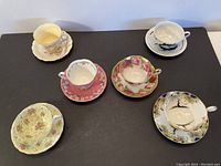 Photo of all six teacups with matching saucers laid on black surface showing variety of floral and decorative patterns.