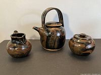 Front view of the studio pottery teapot, creamer, and sugar bowl on a dark surface showing glaze and shapes.
