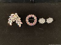 Front view of two Aurora brooches and one pair of clip-on earrings arranged in a row on black background.
