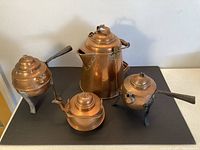 Collection of four copper kettles showing overall shapes and sizes, placed on a black surface against a white wall.