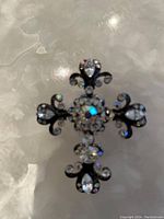 Front view of the cross shaped brooch showing the metal filigree design with rhinestones on the fleur-de-lis ends and center.