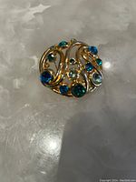 Gold-tone round brooch decorated with blue and green rhinestones photographed on a greyish-white translucent surface.