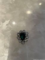 Photo showing the brooch with a green teardrop center stone surrounded by clear round stones on a light surface.