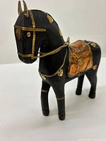 Front angled view of black wooden horse with gold and copper embellishments, detailed saddle, and bridle.