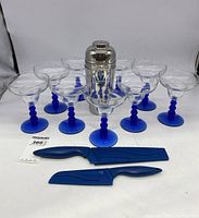 Front view of all items together showing eight margarita glasses, a stainless steel cocktail shaker, and two blue handled knives with sheaths.
