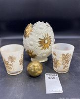 Front view of all items showing ceramic vase with gold floral details, brass tennis ball, and two white Frank Maietta glasses with gold floral patterns.