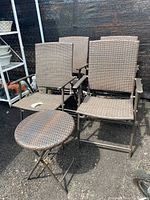 Four outdoor folding chairs with woven synthetic seats and backs and a matching round folding side table with woven top arranged outside on asphalt.