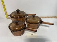 Set of 3 brownish clear Pyrex Vision glass pots with lids, arranged on white surface for scale and reference.